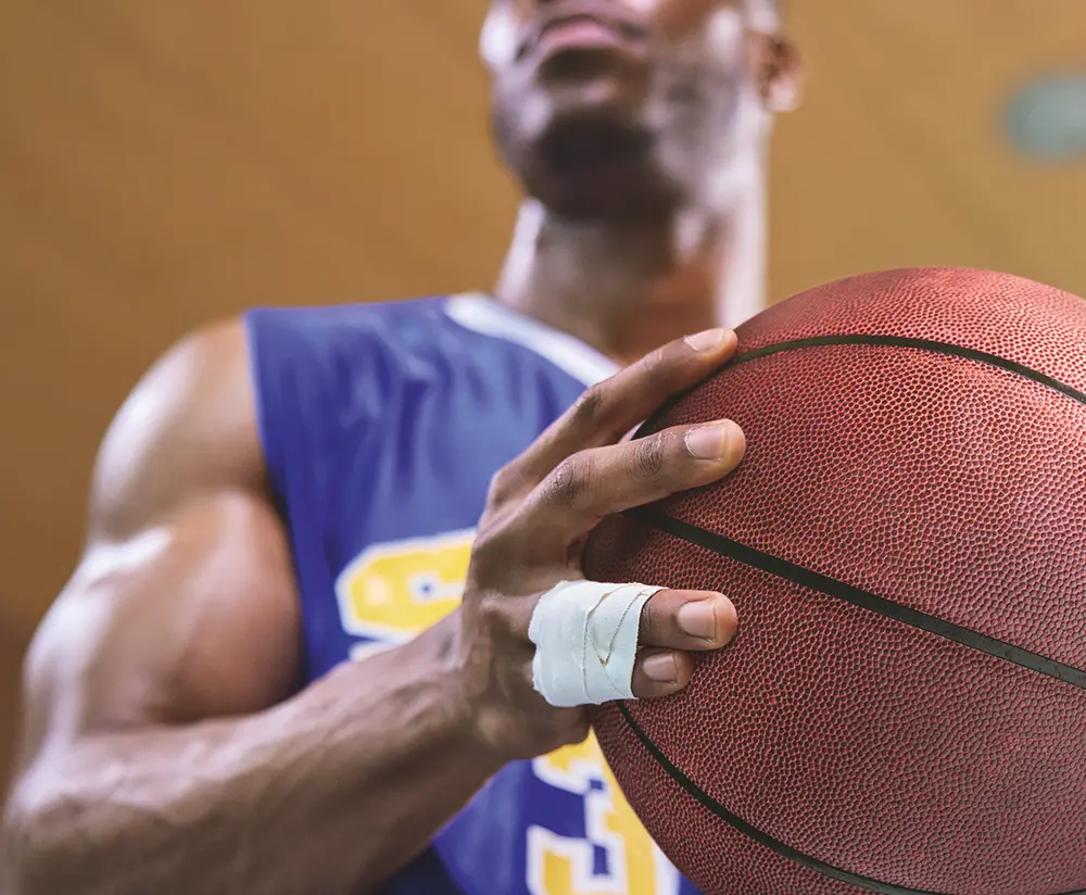 Joueur de basketball tenant un ballon avec un strapping rigide sur deux doigts, illustrant l'utilisation de bandes adhésives rigides pour immobiliser et stabiliser les articulations pendant une activité sportive intense.